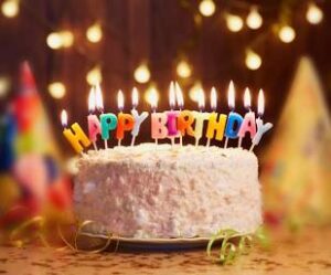 Celebration cake with lit 'Happy Birthday' candles on a plate, colorful party hats in the background.